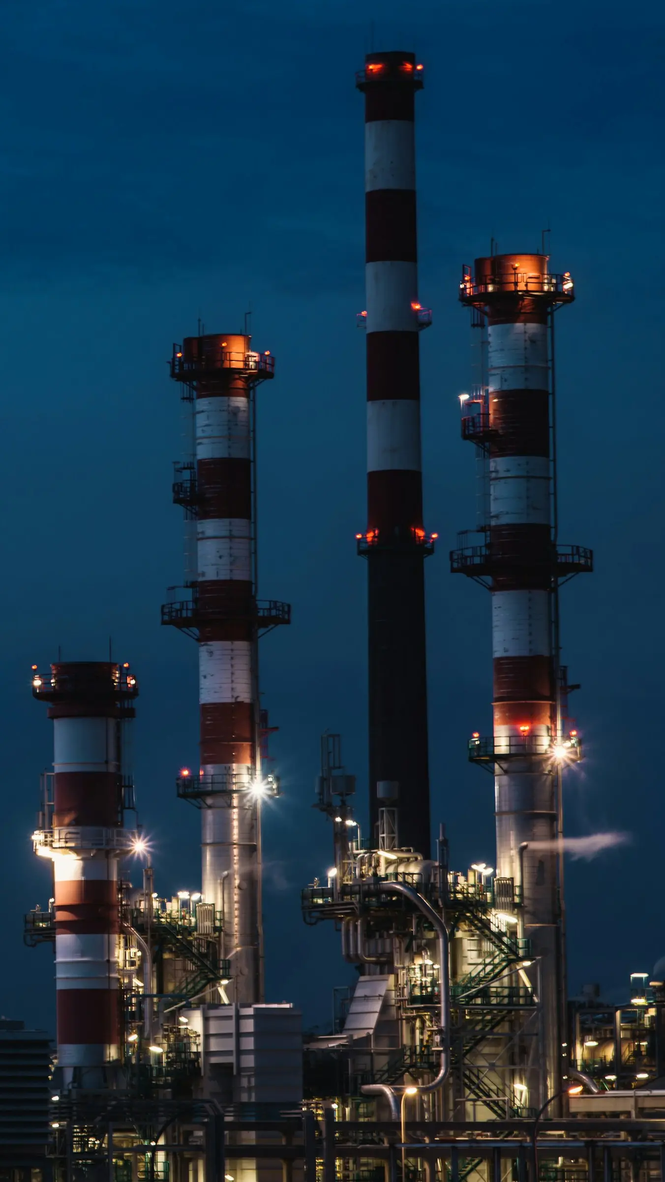 An oil refinery at dusk, its red-and-white stacks lit against a deep blue sky